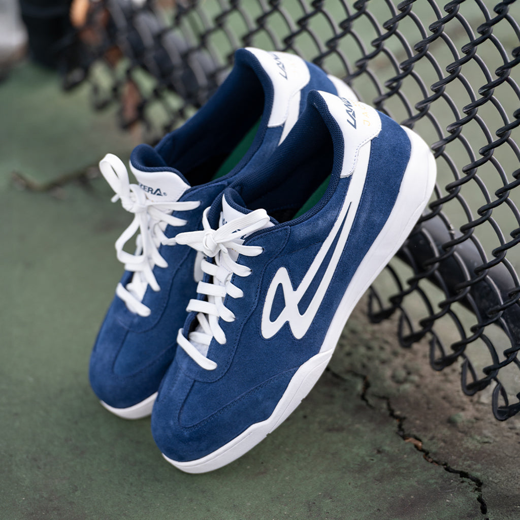 Lanzera Jackal Navy suede sneakers resting against a chain-link fence on an outdoor futsal court, showcasing the rich suede upper, white branding, and non-marking sole for futsal and streetwear performance.